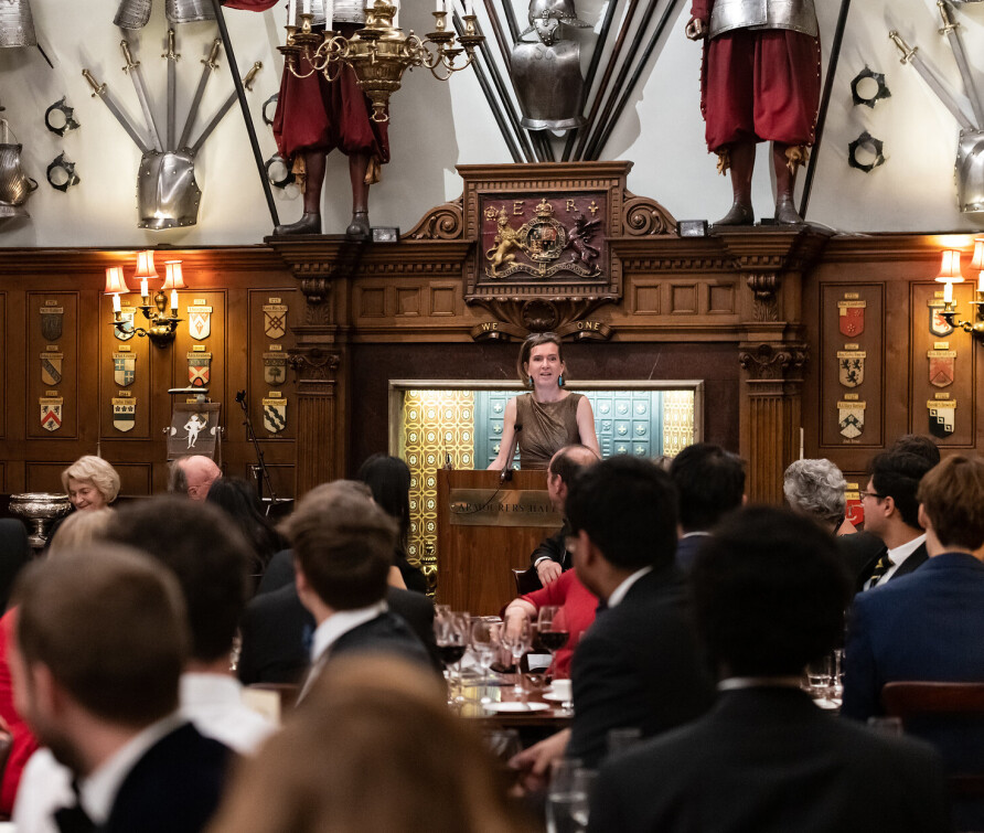 A snapshot from a previous dinner: a female speaker stands at a podium in a grand, old-fashioned hall in front of an audience. The dress code appears to be formal. 