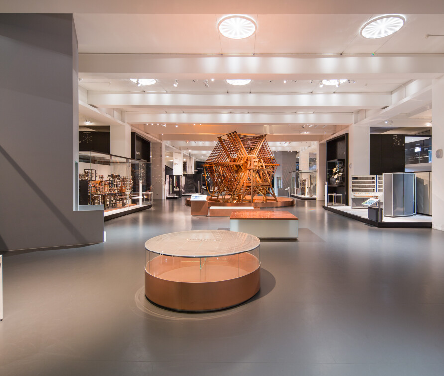 A landscape photo of The Information Age Gallery at the Science Museum, it appears to be spacious, contemporary in its lighting and devoid of people for the photo. Pictured in the centre background is a high copper and wood aerial tuning inductor from Rugby Radio Station. 